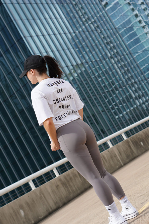 Person wearing a white t-shirt with No Straight Path® text and gray leggings standing against a modern building.