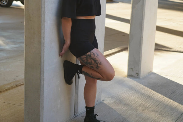 Woman wearing black shorts and shoes leaning against a concrete wall.