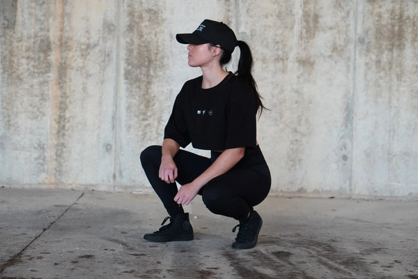Girl in black outfit squatting against a textured wall.