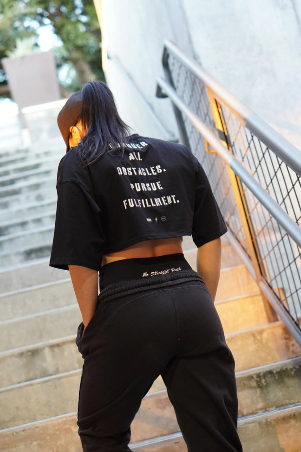 Woman wearing a black t-shirt with text reading 'Conquer all obstacles. Pursue Fulfillment.' on the back, standing outdoors.