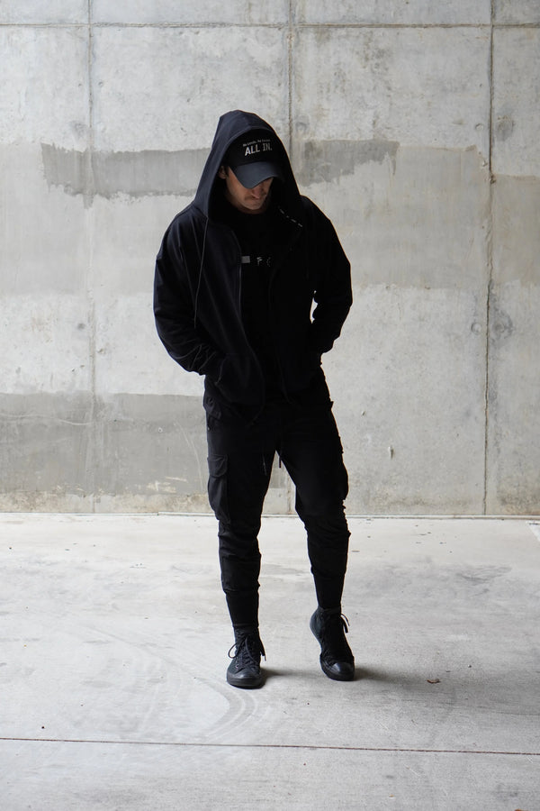 Person wearing a black hoodie and pants standing against a concrete wall.