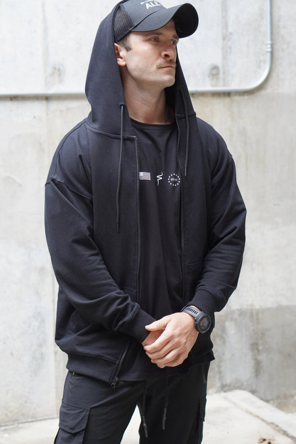 Man wearing a black hoodie and cap with logos, standing against a concrete wall.
