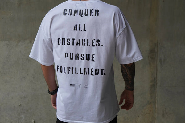 Person wearing a white t-shirt with motivational text reading 'Conquer all obstacles. Pursue fulfillment.' on the back against a concrete wall.