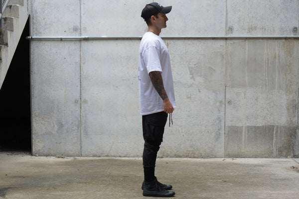Person wearing a white t-shirt, black pants, and a cap standing against a concrete wall.