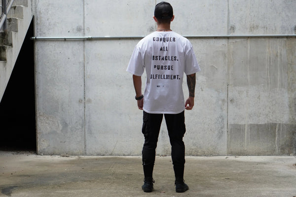 Person wearing a white t-shirt with motivational text reading 'Conquer all obstacles. Pursue Fulfillment.' on the back, standing against a concrete wall.