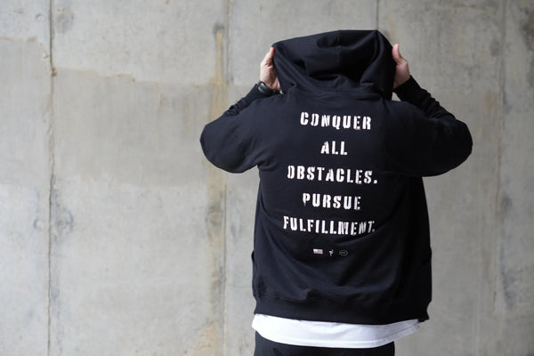 Person wearing a black hoodie with motivational text on the back against a concrete wall.