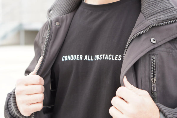 Person wearing a black t-shirt with 'CONQUER ALL OBSTACLES' text, outdoors.