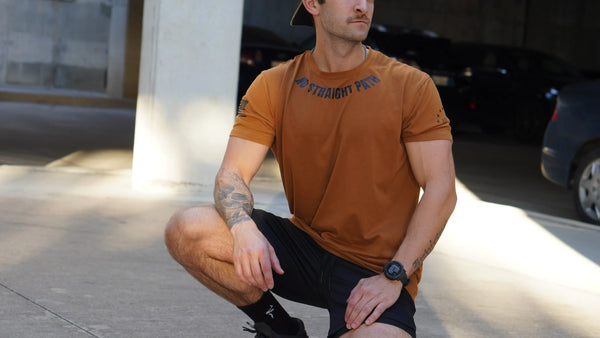 Man in a brown t-shirt and black shorts kneeling outdoors on a sunny day.
