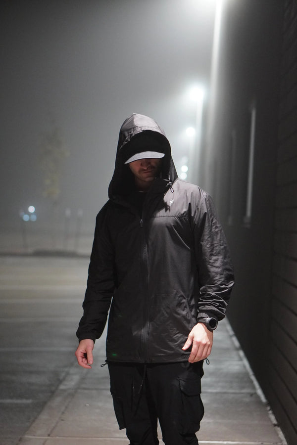 Person wearing a black raincoat with a hood on a foggy street at night.