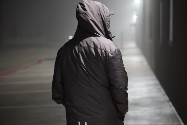 Person wearing a dark hooded jacket on a dimly lit sidewalk under a streetlight.