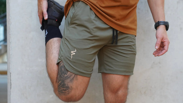 Person wearing green shorts with a white No Straight Path® logo, standing against a concrete wall.