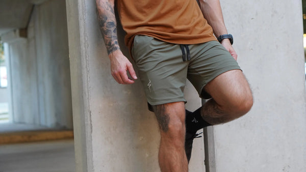 Person wearing a tan shirt and green shorts leaning against a concrete wall.