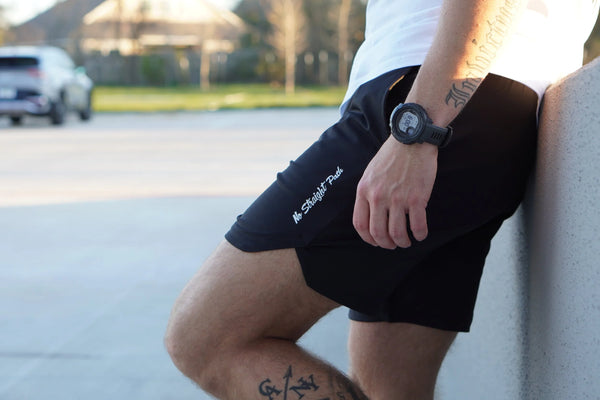 Person wearing black shorts with No Straight Path® text, sitting on a curb outdoors.