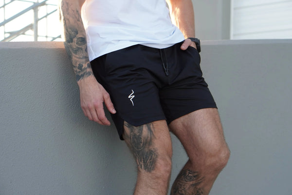 Person wearing black shorts with a No Straight Path® logo, standing against a gray wall.