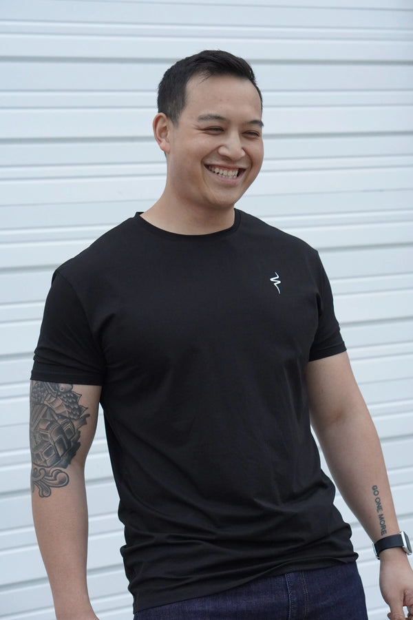 Man smiling wearing a black t-shirt with a No Straight Path® logo, standing against a white wall.