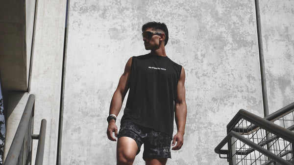 Man wearing a black sleeveless shirt and shorts walking outdoors against a concrete wall.