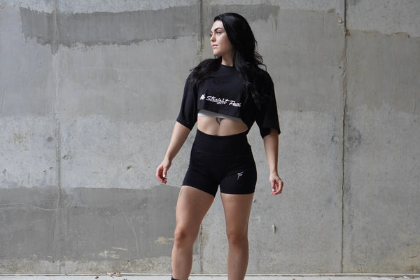 Woman wearing a black crop top and shorts with visible branding against a concrete wall.