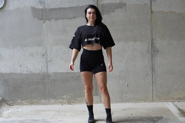 Woman wearing a black crop top and shorts standing against a concrete wall.