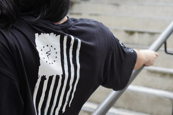 Person wearing a black t-shirt with a white flag design on the back, standing on steps.