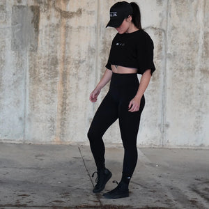 Woman wearing a black cropped shirt and leggings standing against a textured wall.