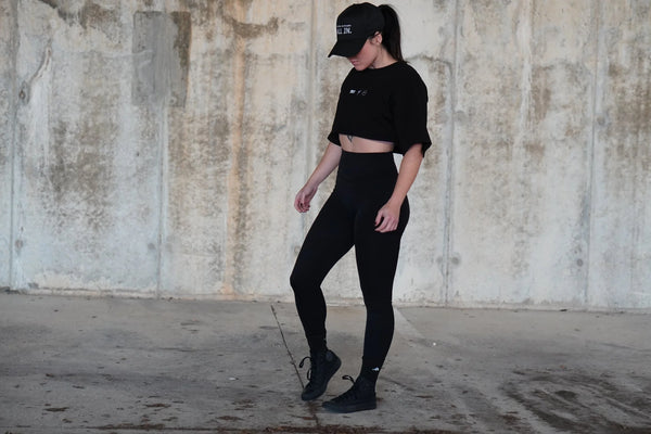 Woman wearing a black cropped shirt and leggings standing against a textured wall.
