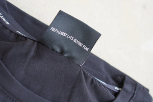 Black shirt with a tag that reads 'Fulfillment Lies Beyond Fear' on a neutral background