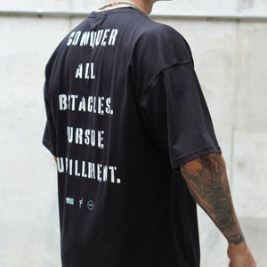 Person wearing a black t-shirt with text on the back, standing against a light-colored wall.