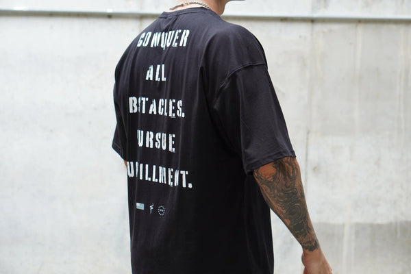 Person wearing a black t-shirt with text on the back, standing against a light-colored wall.