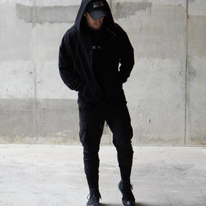 Person wearing a black hoodie and pants standing against a concrete wall.