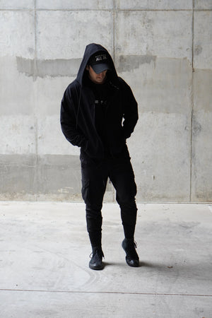 Person wearing a black hoodie and pants standing against a concrete wall.