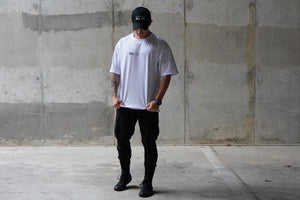 Person wearing a white t-shirt and black pants standing against a concrete wall.