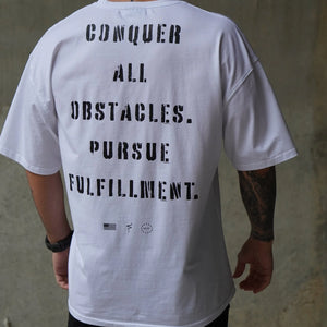Person wearing a white t-shirt with motivational text reading 'Conquer all obstacles. Pursue fulfillment.' on the back against a concrete wall.