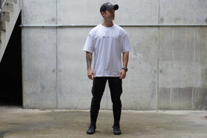 Man wearing a white t-shirt and black pants standing against a concrete wall.