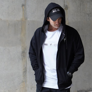 Person wearing a black hoodie and cap with a white shirt, standing against a concrete wall.