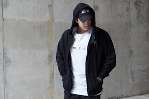 Person wearing a black hoodie and cap with a white shirt, standing against a concrete wall.