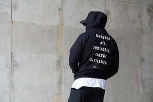 Person wearing a black hoodie with text on the back against a concrete wall.