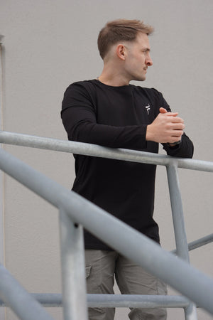 Man wearing a black long-sleeve shirt and beige pants standing against a plain wall.
