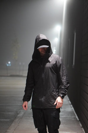 Person wearing a black raincoat with a hood on a foggy street at night.