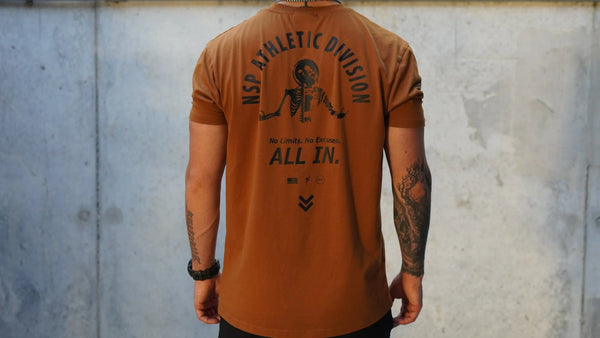Person wearing a brown t-shirt with text and graphics on the back against a concrete wall.