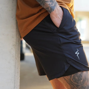 Person wearing black shorts with a No Straight Path® logo, leaning against a concrete wall.