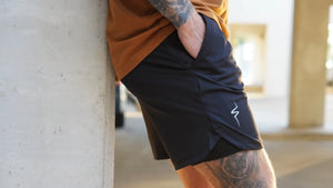 Person wearing black shorts with a No Straight Path® logo, leaning against a concrete wall.