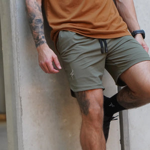 Person wearing a tan shirt and green shorts leaning against a concrete wall.