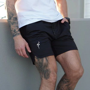 Person wearing black shorts with a No Straight Path® logo, standing against a gray wall.