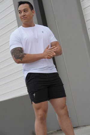 Man wearing a white t-shirt and black shorts with tattoos on his arm, standing against a plain background.