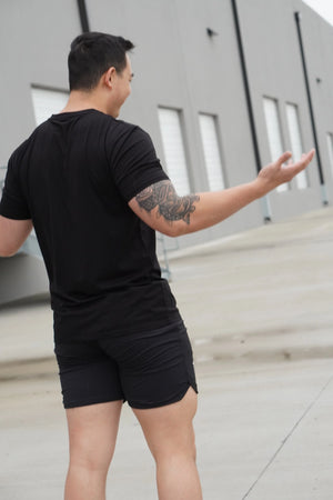 Man in black t-shirt and shorts standing outdoors with arms outstretched