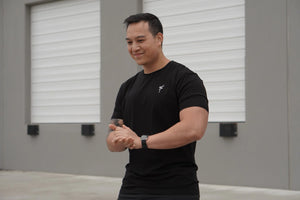 Man wearing a black t-shirt with a white No Straight Path® logo, standing indoors with gray walls and windows.