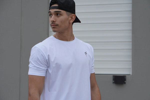 Man wearing a white t-shirt with a small No Straight Path® logo and a black cap, standing against a plain background.