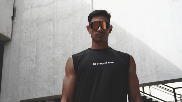 Man wearing a black sleeveless shirt with 'No Straight Path®' text, standing against a concrete wall.