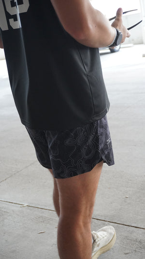 Person wearing black shorts with white No Straight Path® logo on a light gray pavement.