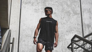 Man wearing a black sleeveless shirt and shorts walking outdoors against a concrete wall.